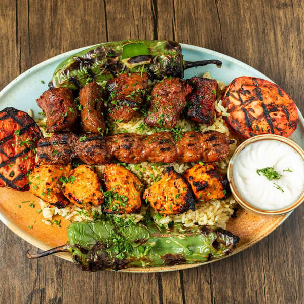 A Mediterranean platter with various grilled meats, rice, charred vegetables, and a side of white sauce.
