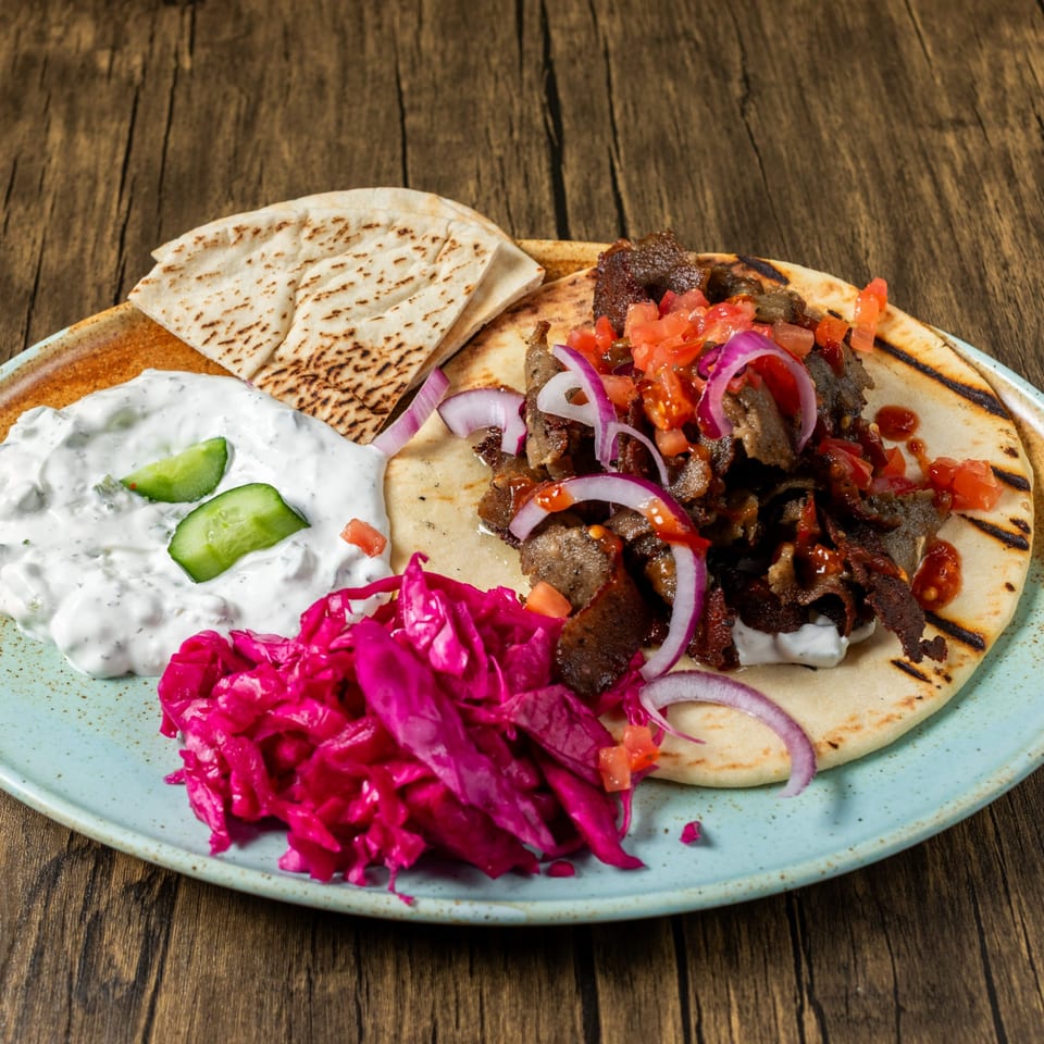 Mediterranean gyro plate with meat, red onions, tomatoes, tzatziki, pickled cabbage, and pita bread.
