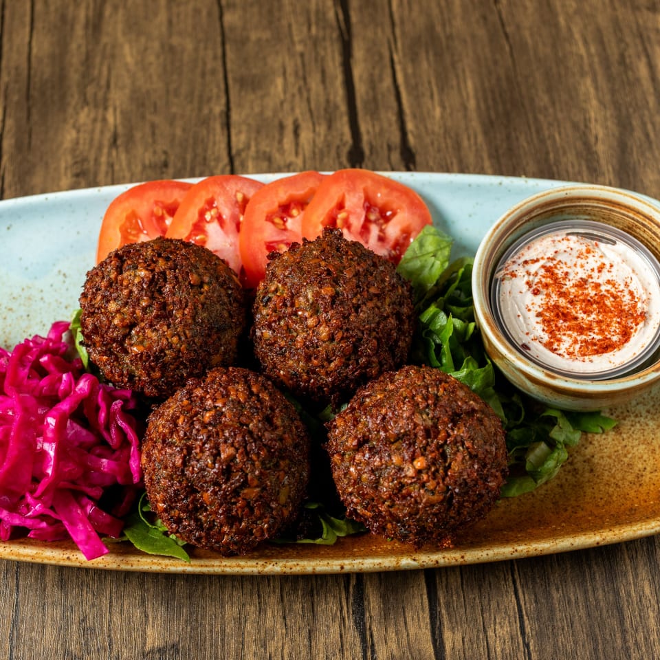 Four falafel balls on a bed of lettuce with sliced tomatoes, pickled cabbage, and a dip.