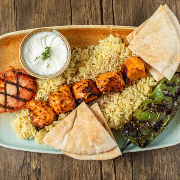 Mediterranean chicken kebab with rice, grilled tomato, pepper, pita bread, and a side of yogurt sauce.