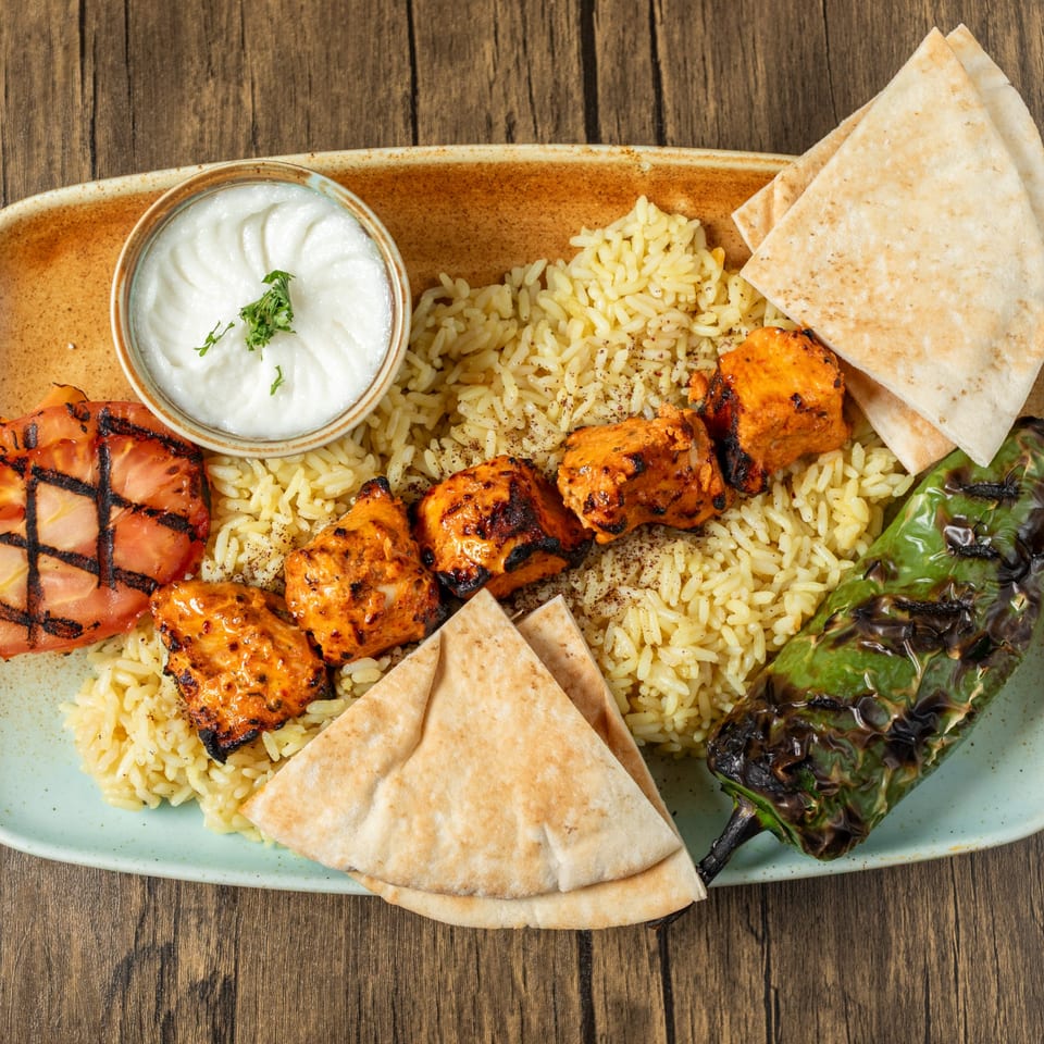 Mediterranean chicken kebab with rice, grilled tomato, pepper, pita bread, and a side of yogurt sauce.